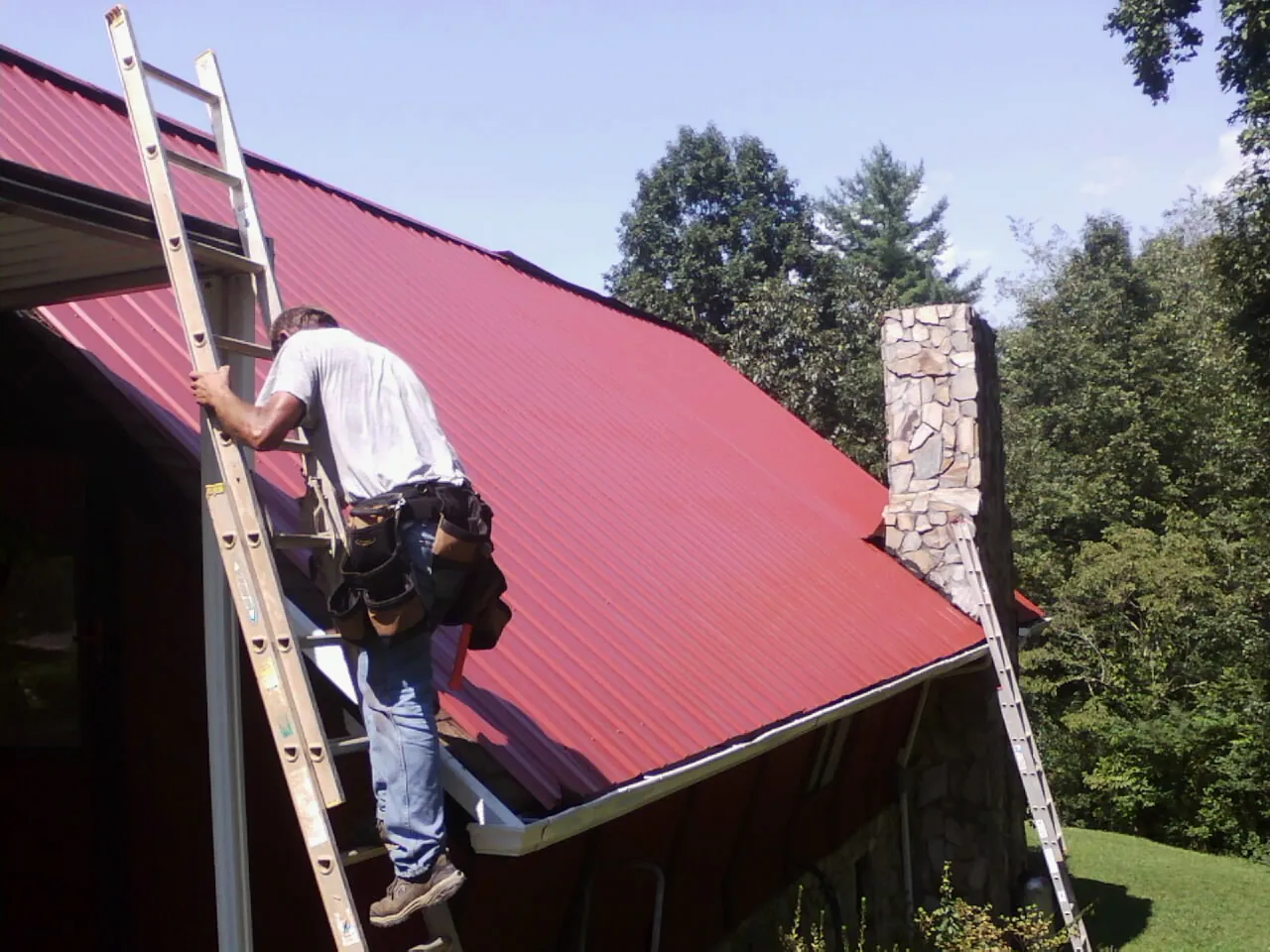 Roofer on a ladder beside a red metal roof and stone chimney — Soffit & Fascia Repair by Roofing Brothers in Abington